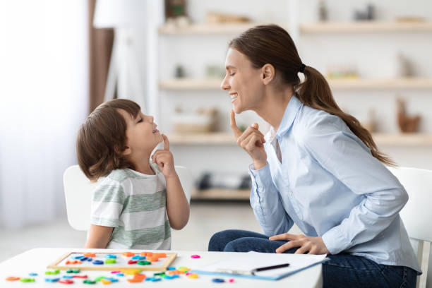 istockphoto-1387959076-612×612 Speech training for kids. Professional woman specialist training with little boy at cabinet, teaching him right articulation exercises, side view
